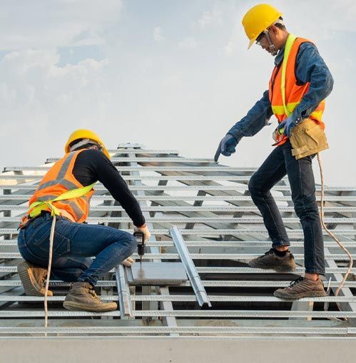 Two construction workers on a roof, wearing safety vests and hard hats.