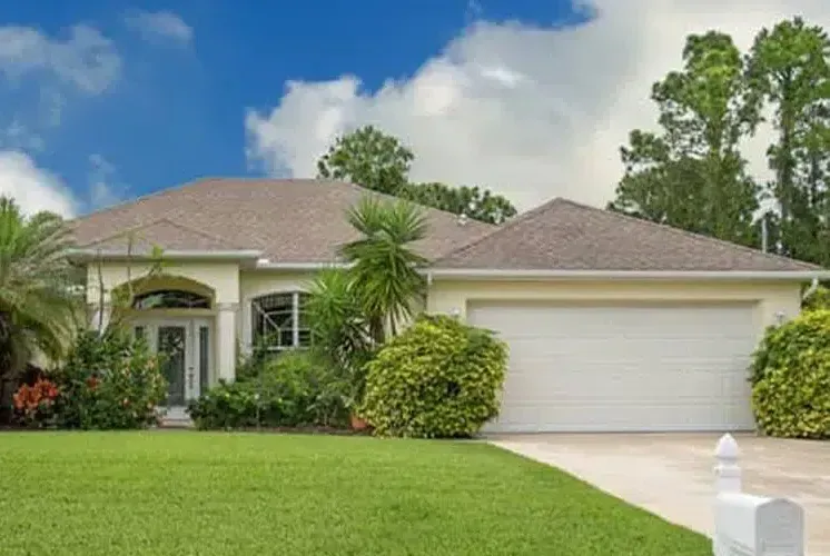 Yellow house with a two-car garage, lush green lawn, and blue sky with clouds.