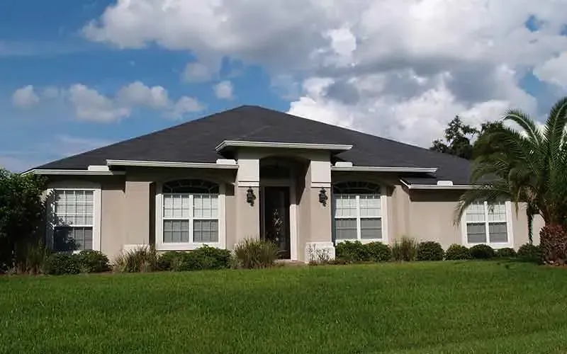 Single-story stucco house with a dark roof and green lawn under a cloudy sky.