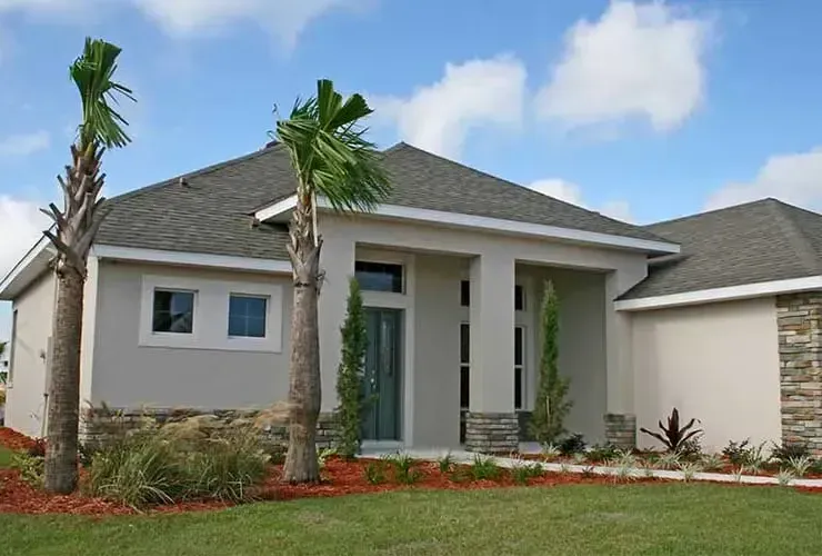 Light gray house with palm trees, stone accents, and green grass under a blue sky with puffy clouds.