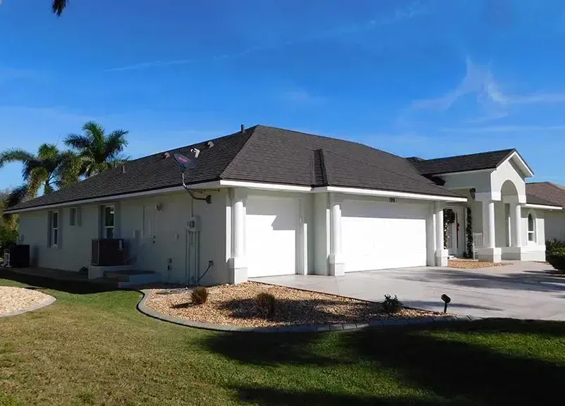 White stucco house with a black roof, two-car garage, and green lawn on a sunny day.