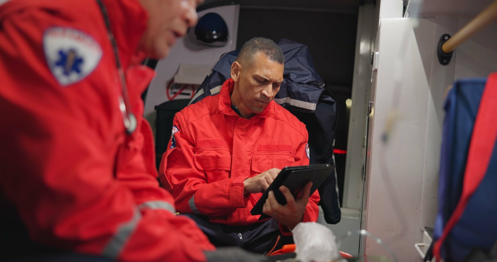 Two emergency medical personnel in red jackets inside an ambulance; one uses a tablet.