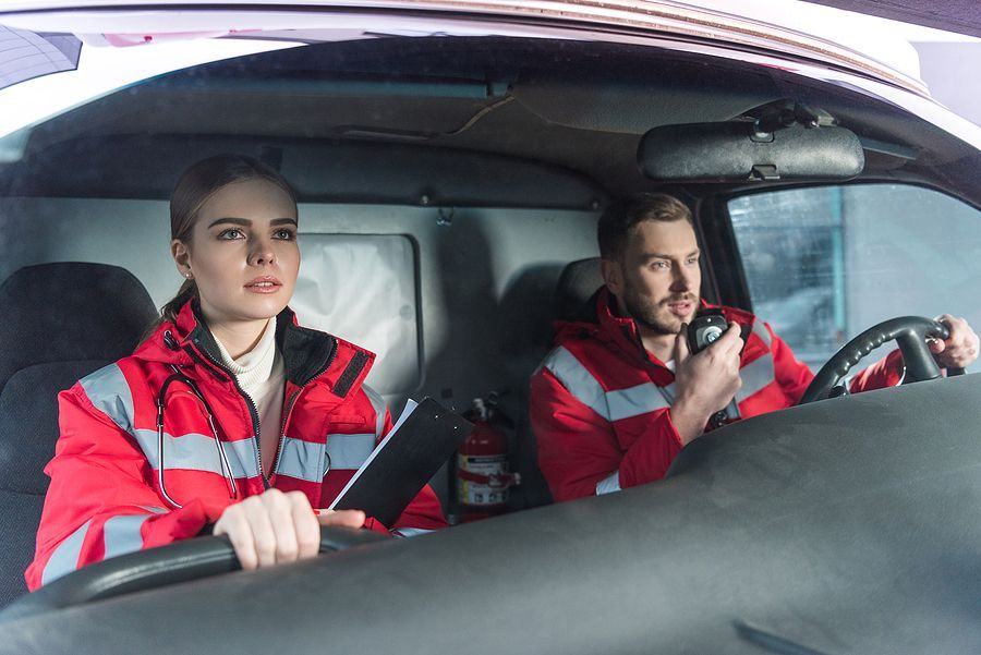 Two paramedics in red jackets inside an ambulance. The driver speaks into a radio, the other looks forward with a clipboard.