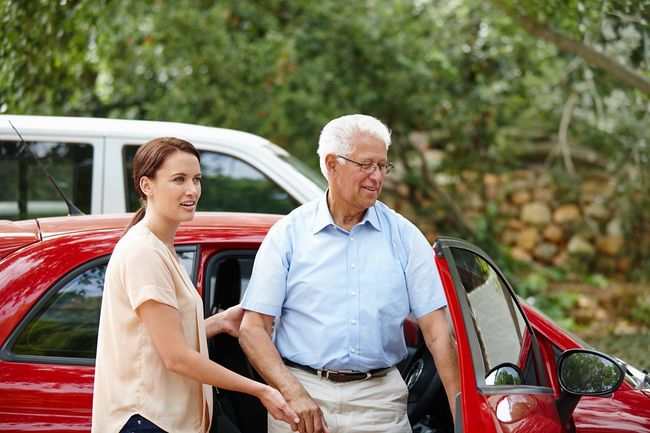 Woman helping a man exit a red car, outdoors. Man holds car door, woman extends hand.