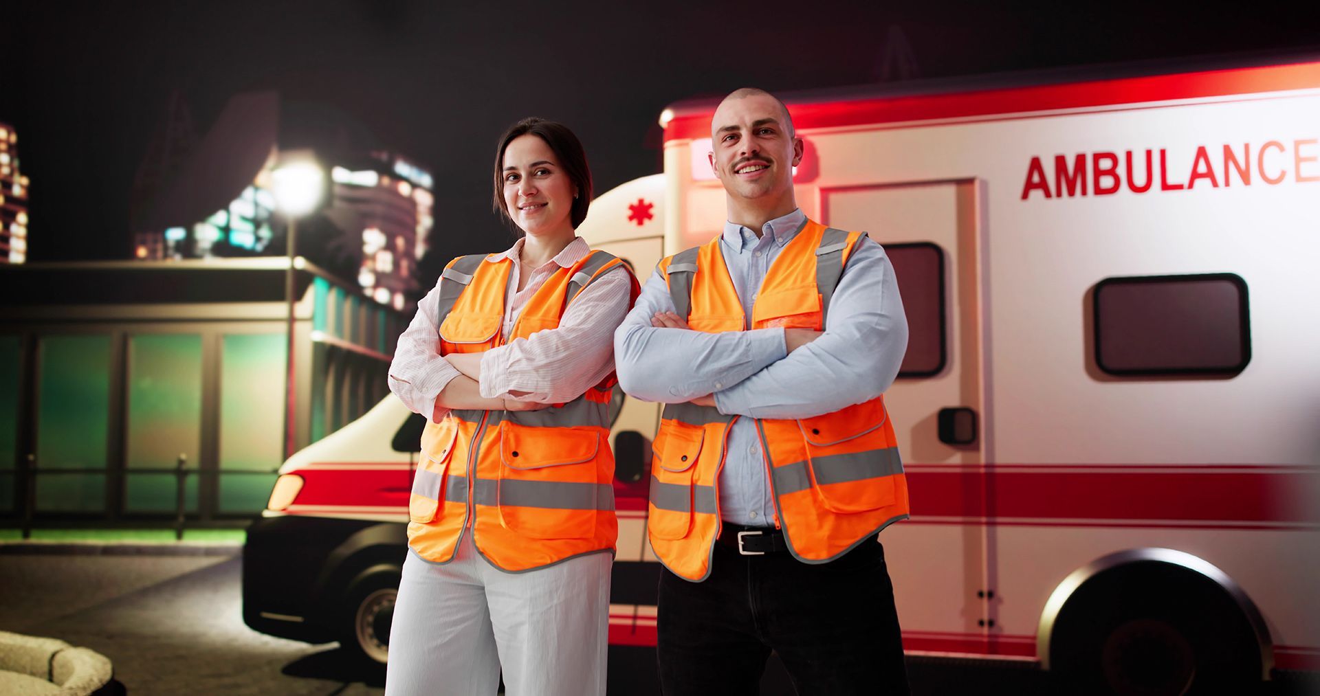 Two people in orange vests stand in front of an ambulance at night. Both have arms crossed, smiling.