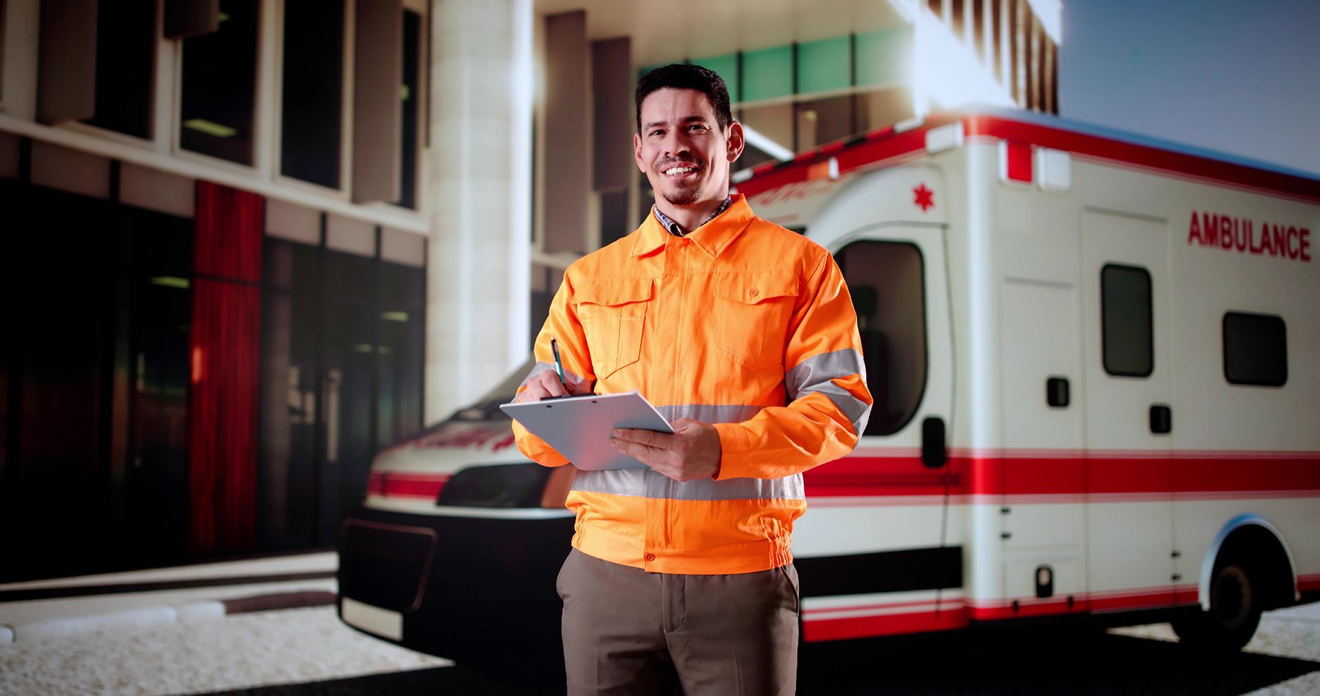 Smiling man in orange vest holding clipboard stands in front of an ambulance near a building.