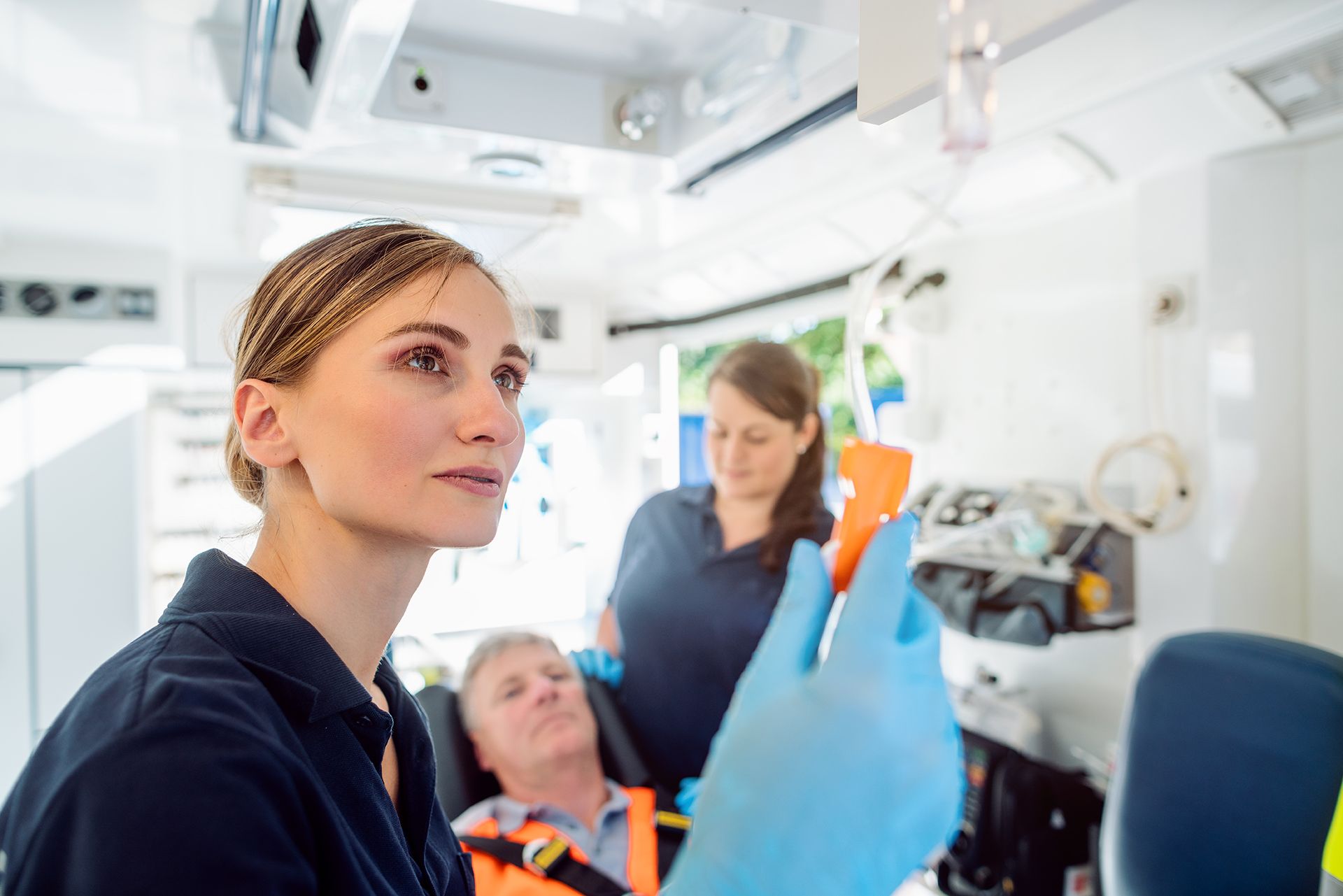 EMT tending to a patient in an ambulance, another EMT in the background. Blue gloves and uniforms.