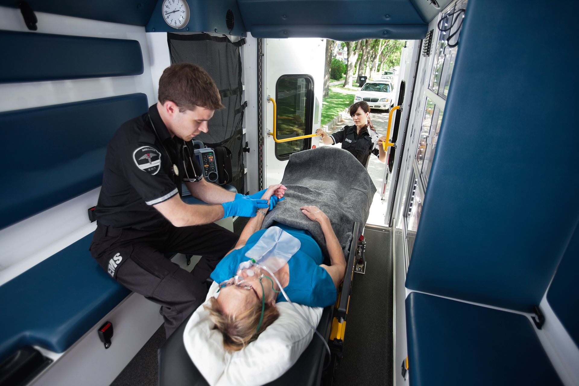 Inside an ambulance, EMT attends to a patient on a stretcher, another EMT stands in doorway.