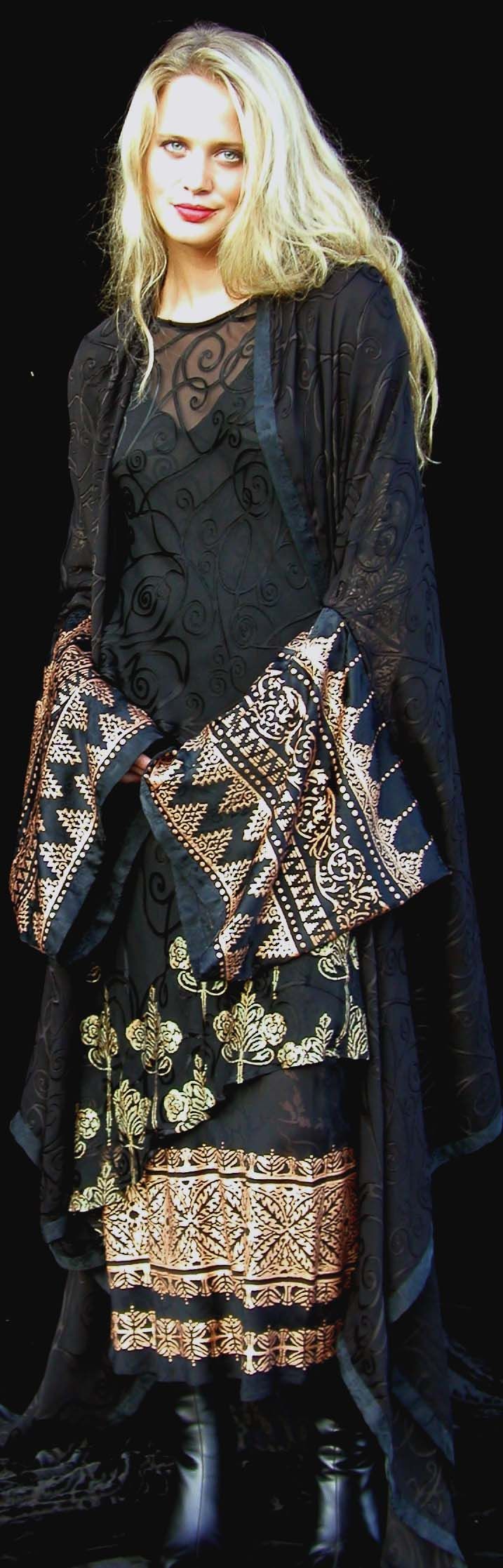 Woman in black dress with gold embellishments, long blonde hair, standing in front of black background.