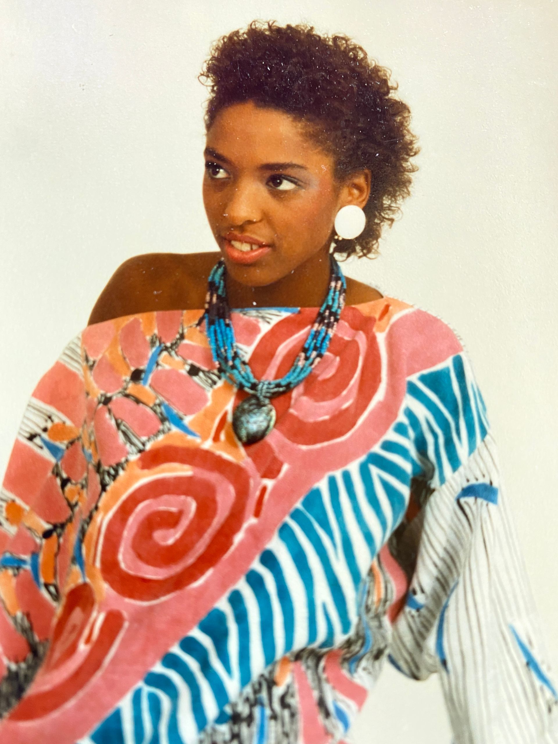 Woman in colorful patterned top and blue necklace, looking off-camera, with short curly hair and white earrings.