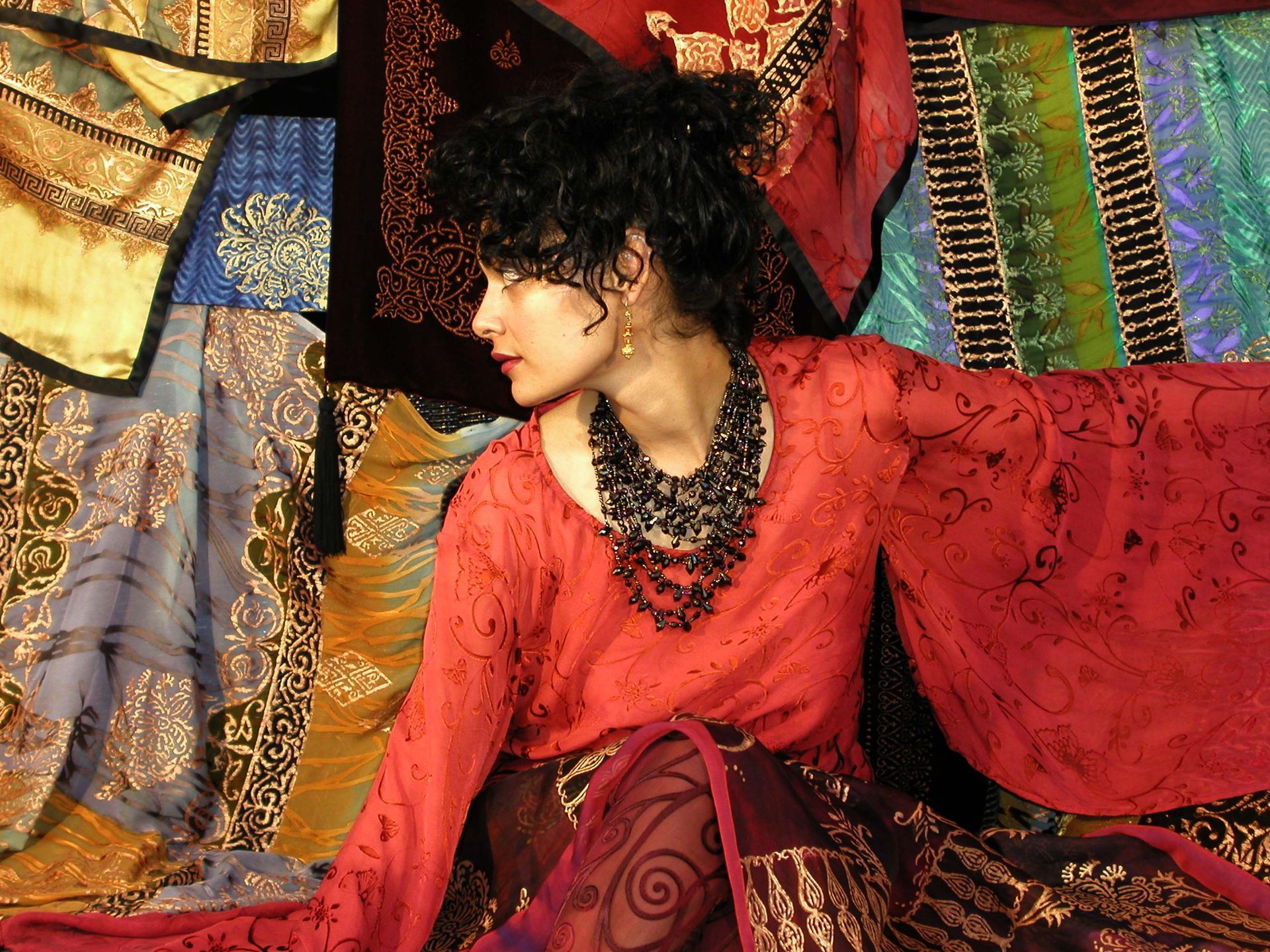 Woman in red top with dark necklace, posing in front of colorful patterned fabric.