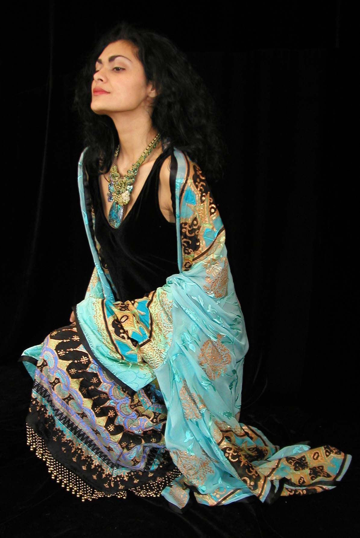 Woman kneels on black surface, wearing blue, gold, and black patterned fabric over black dress.