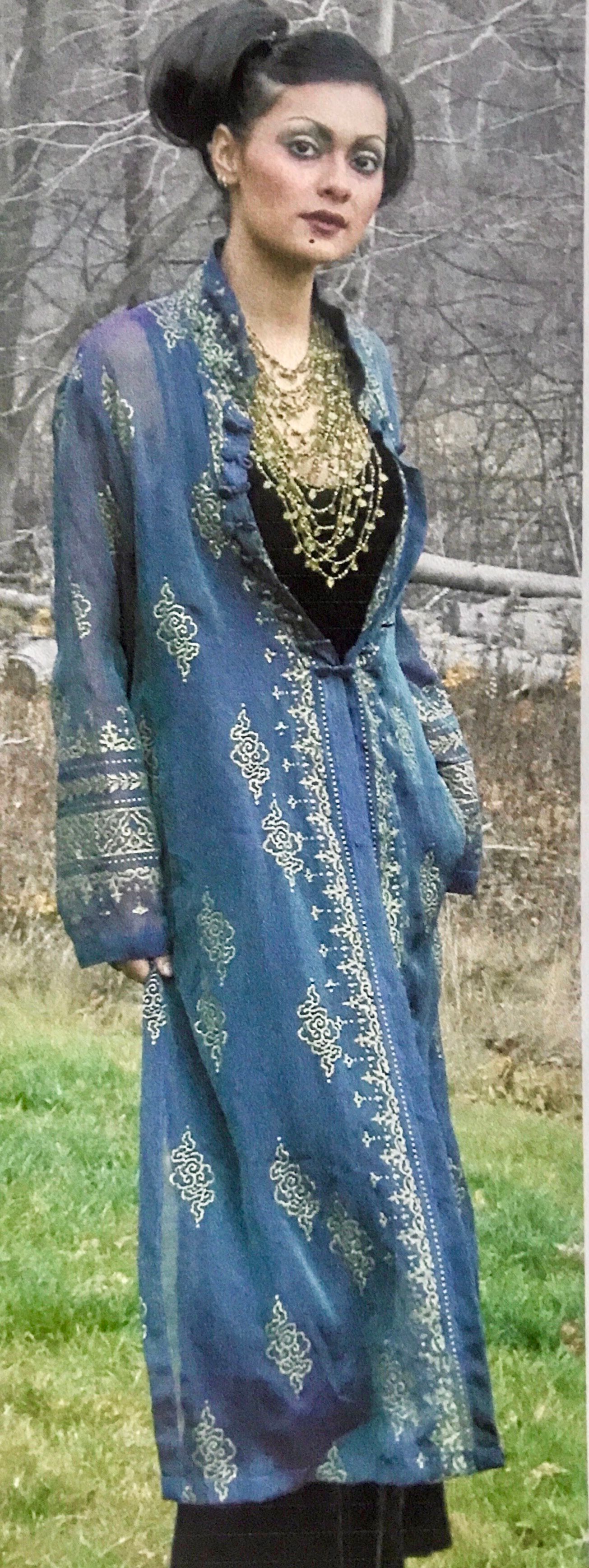 Woman in blue patterned robe with gold necklace, standing in front of greenery.