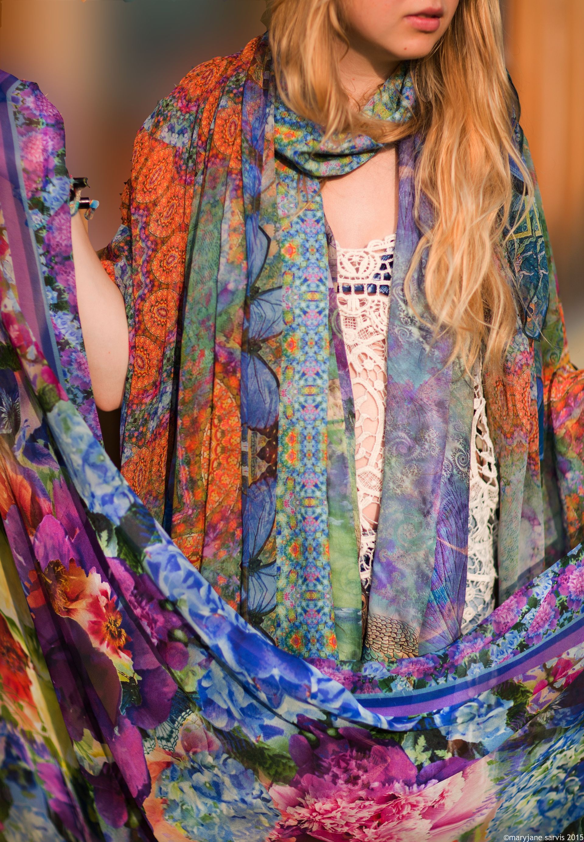 Woman wearing a colorful patterned scarf and a white lace top, holding up fabric with floral and geometric designs.