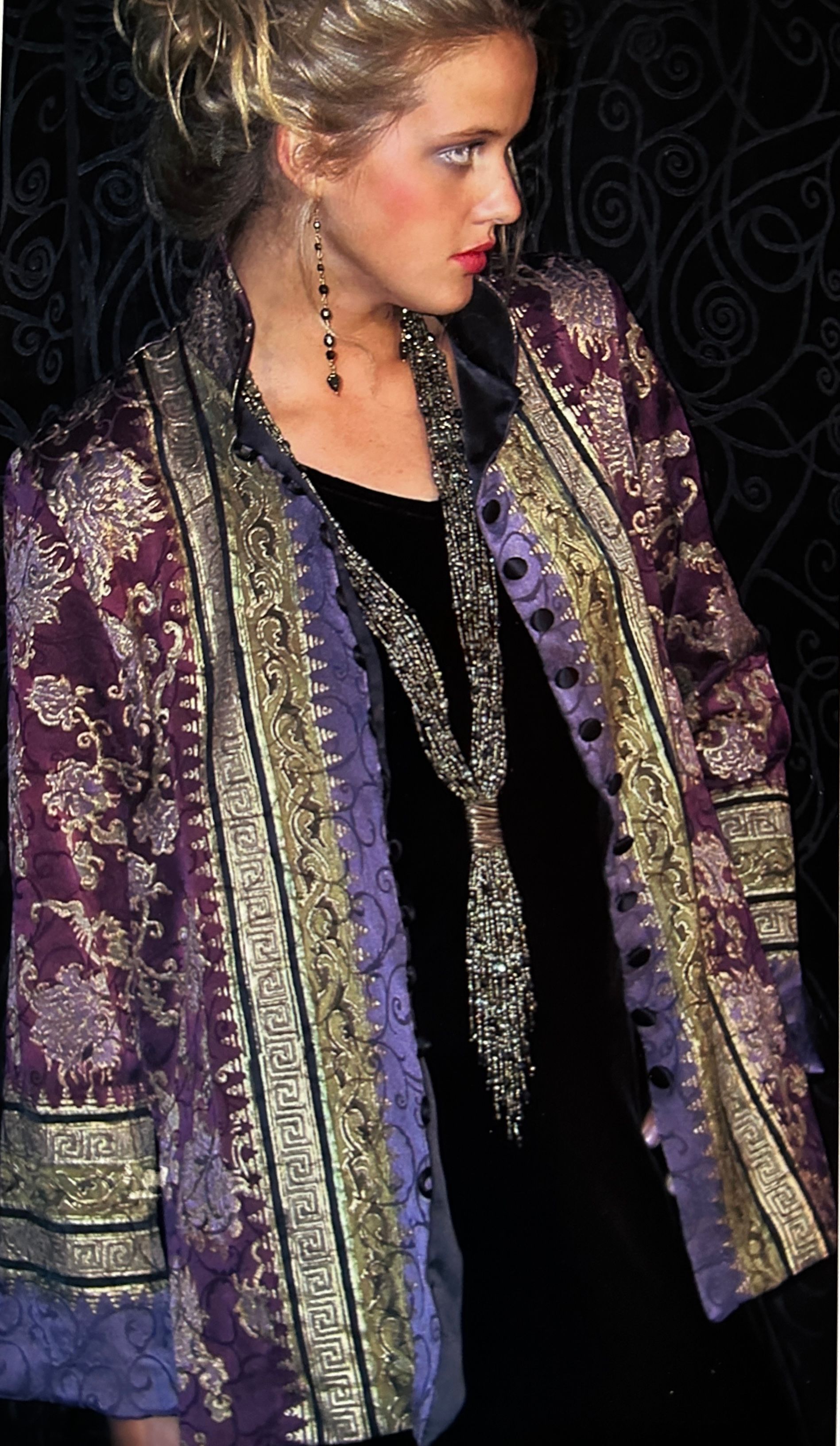 Woman wearing a purple, gold and black patterned jacket and long beaded necklace.