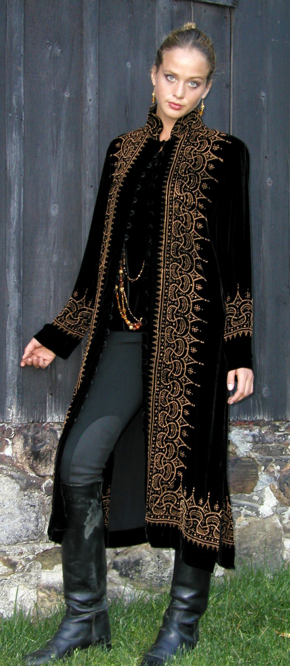 Woman in black coat with gold embroidery, black pants, and boots, standing outdoors.