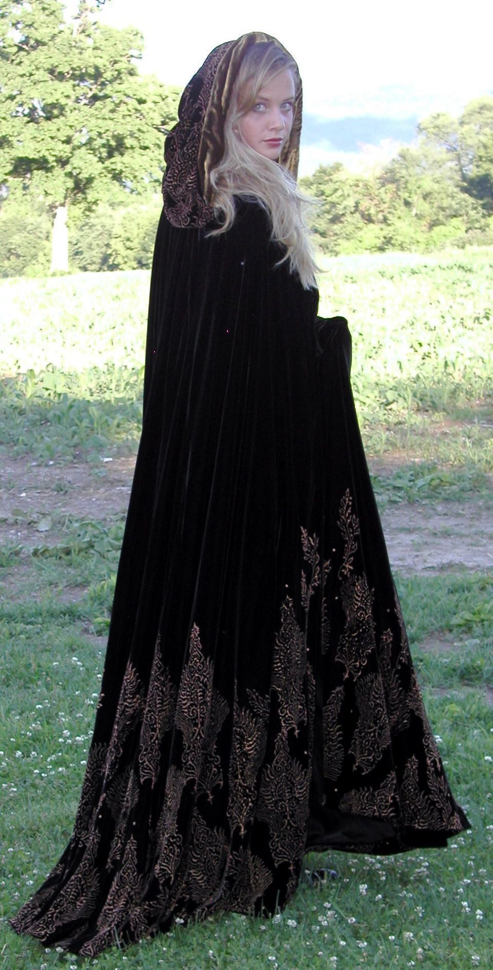 Woman in a black velvet cloak with gold embroidery, standing outdoors.