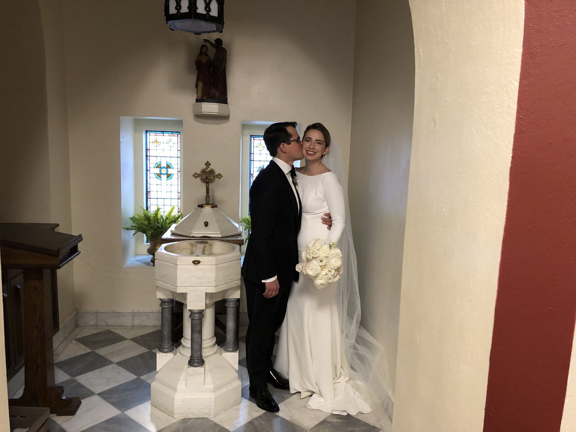 Newlyweds in a church hallway. The groom kisses the bride's cheek. She holds a bouquet, he wears a black suit.