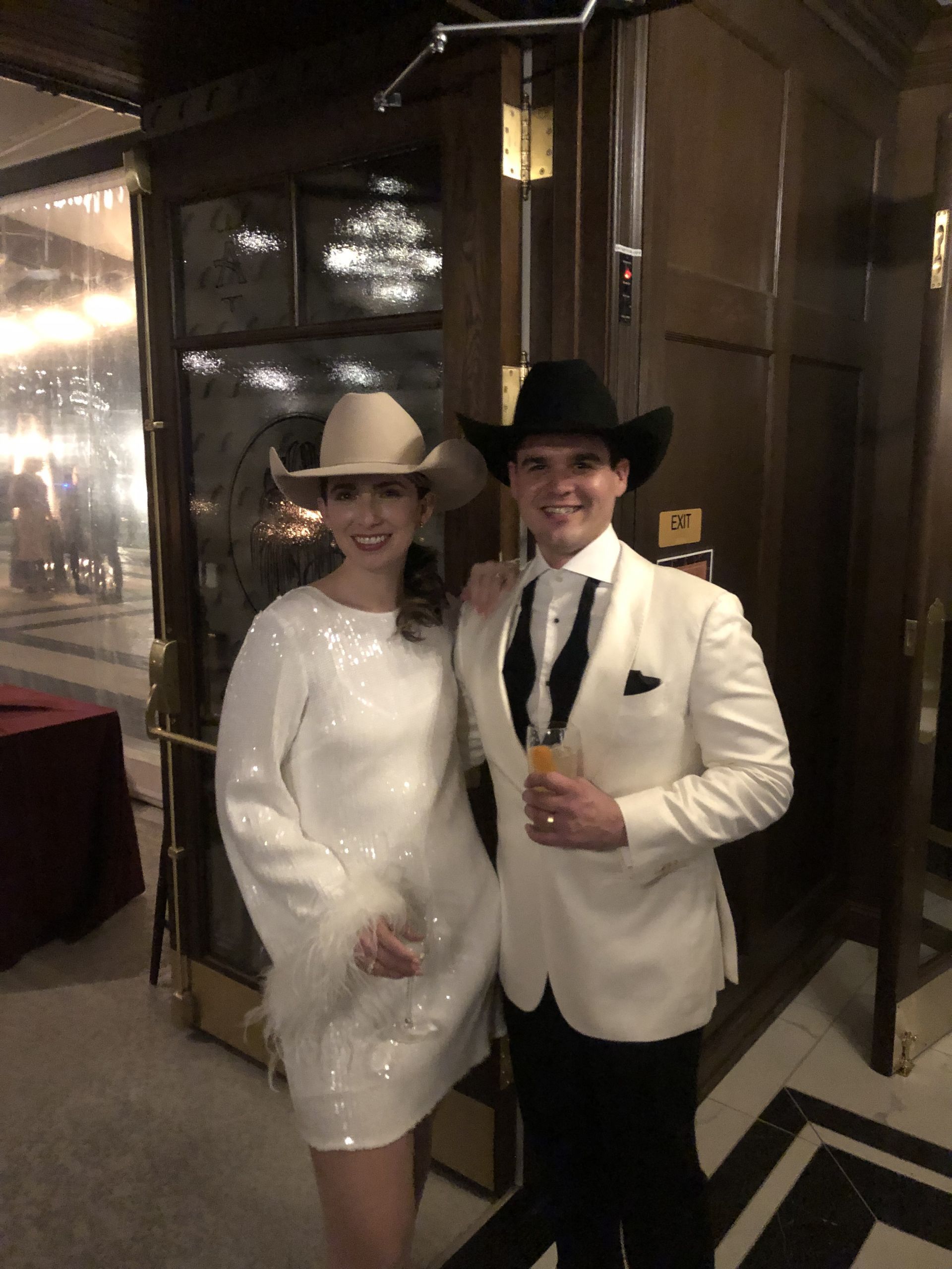 Two people in formal cowboy attire pose by a door. The woman wears a white dress and hat; the man, a white suit and black hat.