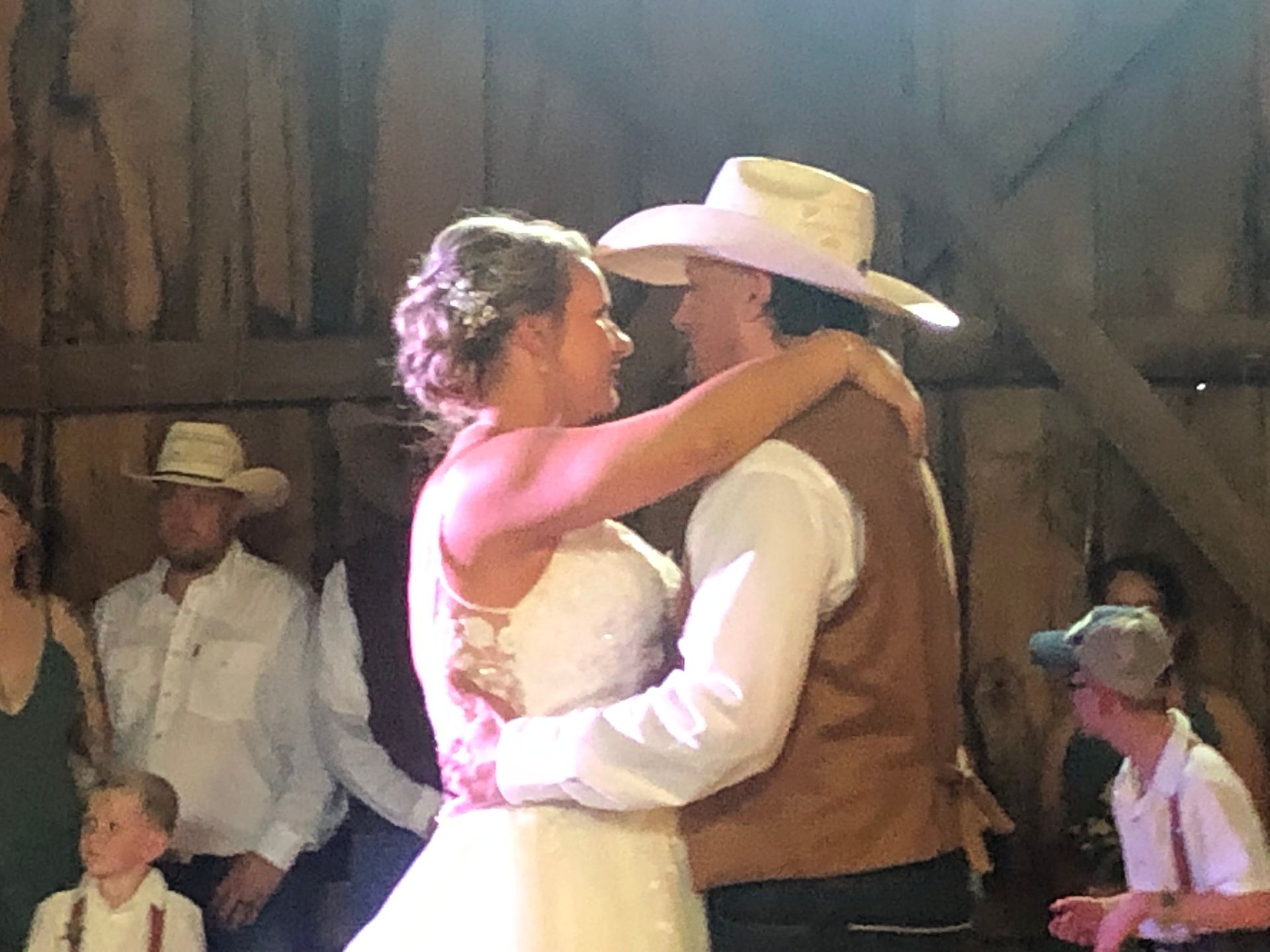 Bride and groom dance in barn, both wearing cowboy hats, surrounded by wedding guests.