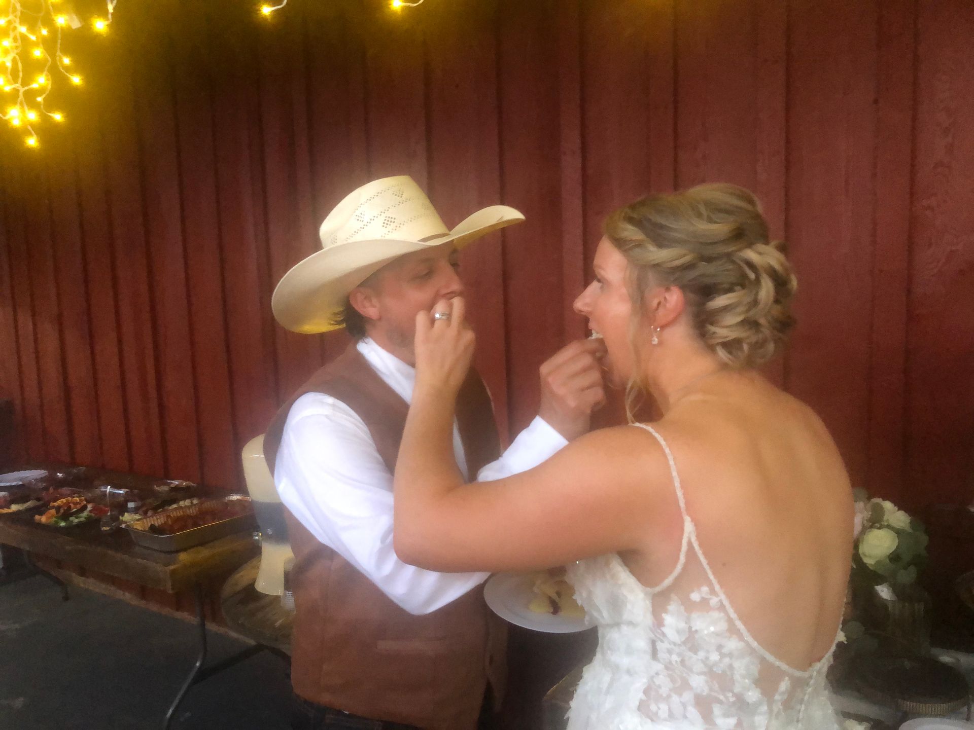 Bride and groom feed each other cake at outdoor wedding, against a wooden barn backdrop.
