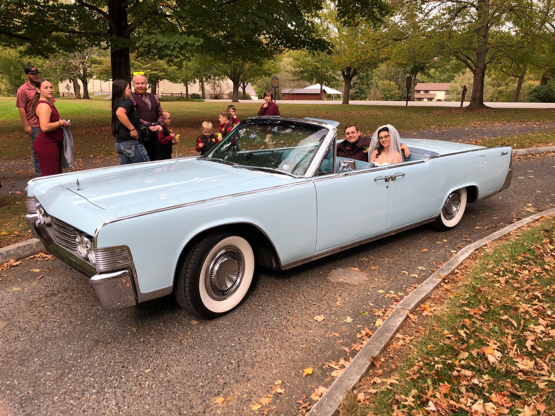 Newlyweds pose by a light blue vintage convertible. The man wears a burgundy suit; the woman, a white dress and jean jacket.