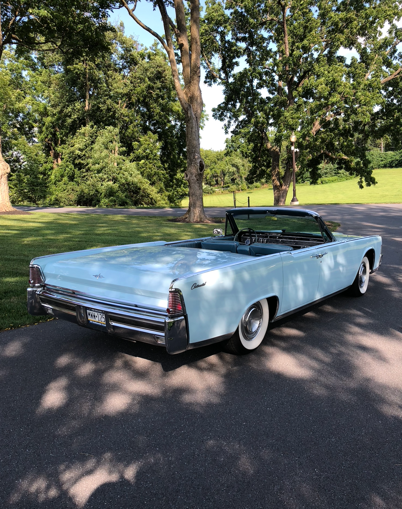 Light blue vintage convertible car with white-wall tires, parked on asphalt, trees in the background.