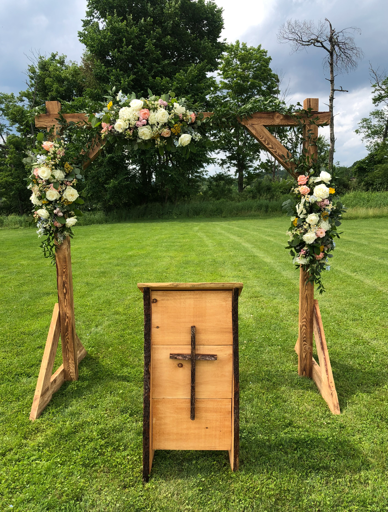 Wooden wedding arch decorated with flowers on a grassy field, with a wooden podium in front.