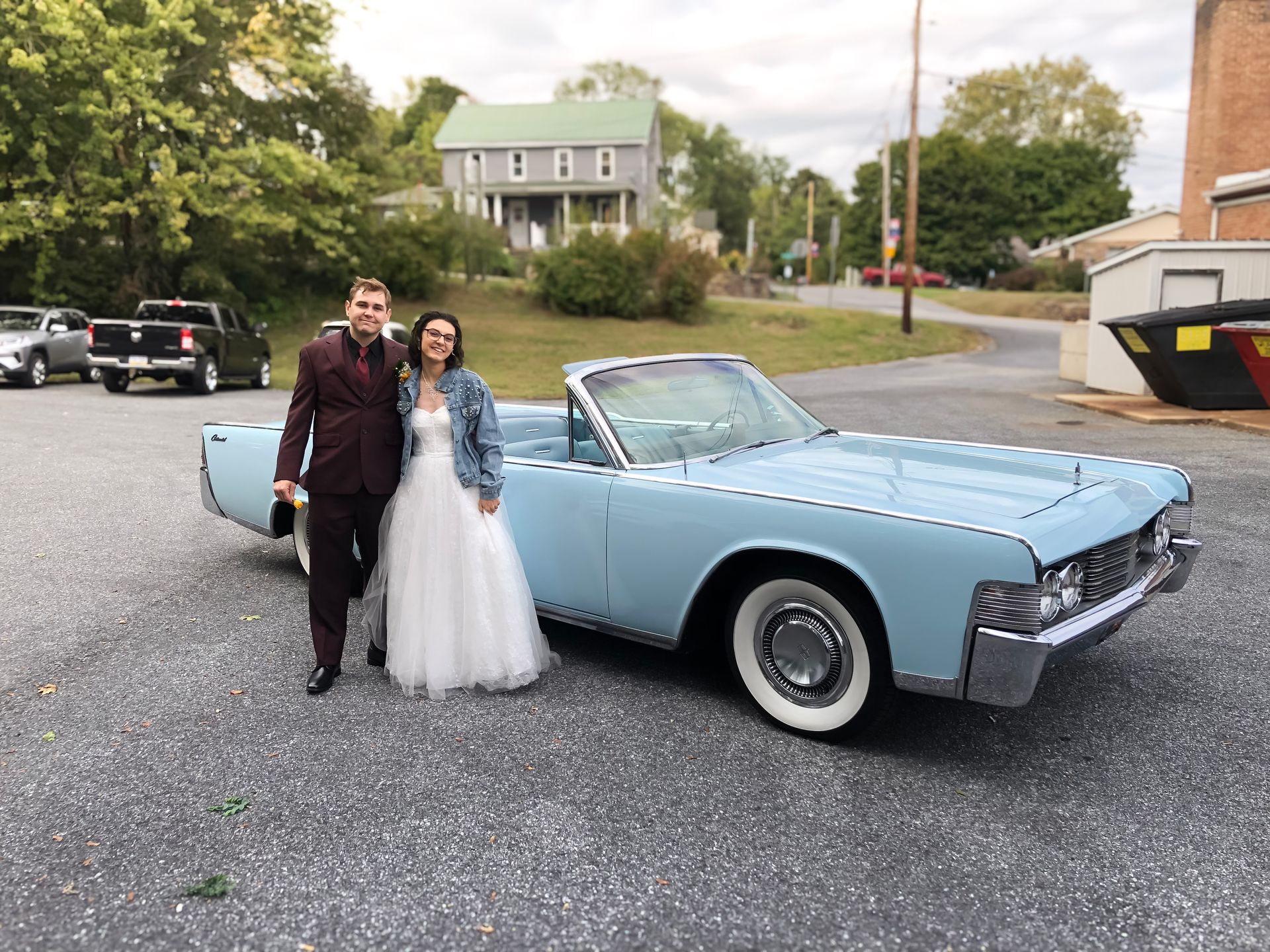 Couple poses beside a light blue convertible, next to a building in a town, one wearing a suit and the other a wedding dress.