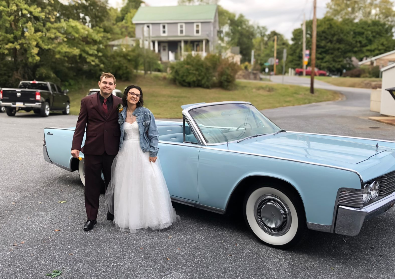 Newlyweds pose by a light blue vintage convertible. The man wears a burgundy suit; the woman, a white dress and jean jacket.