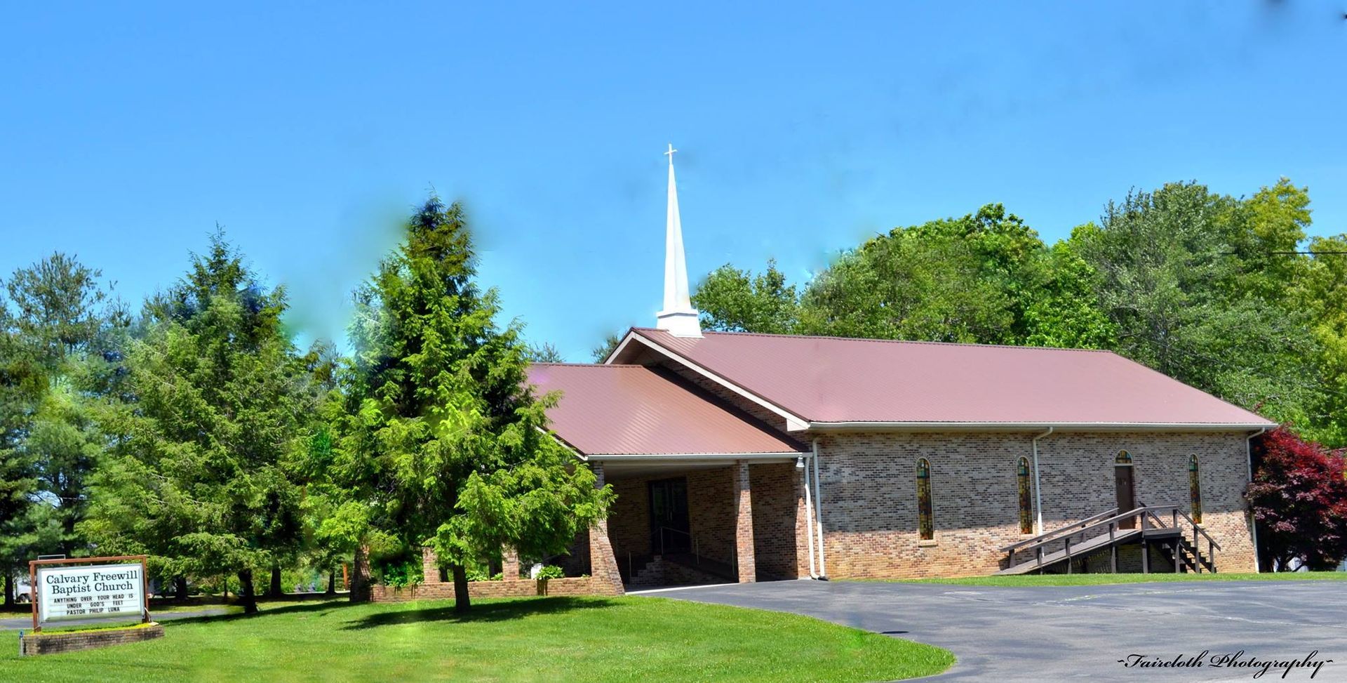 Church building with brown metal roof, brick facade, steeple, and trees.