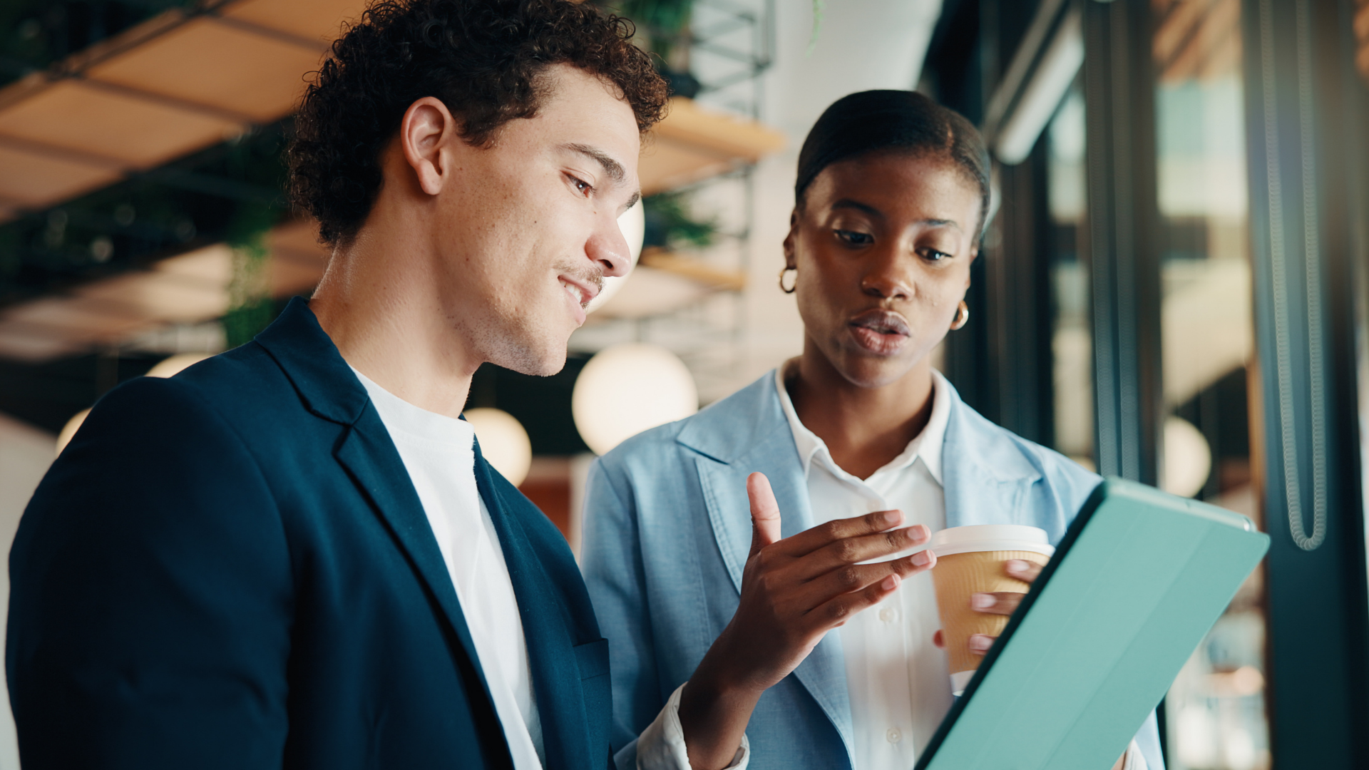 Two coworkers review a tablet together in a bright office, one pointing while the other looks on.