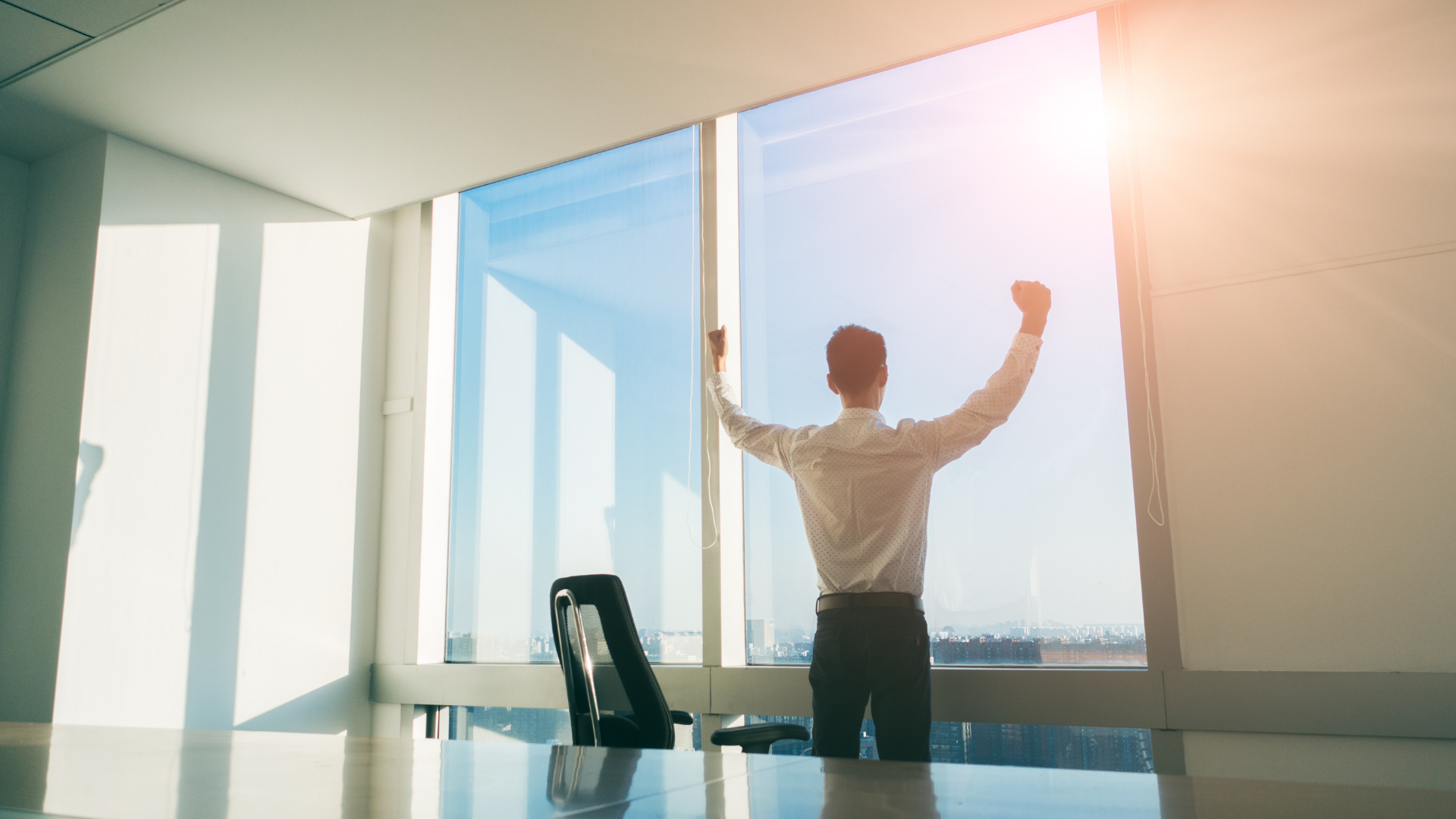 Person celebrating in a bright office with arms raised beside floor-to-ceiling windows.