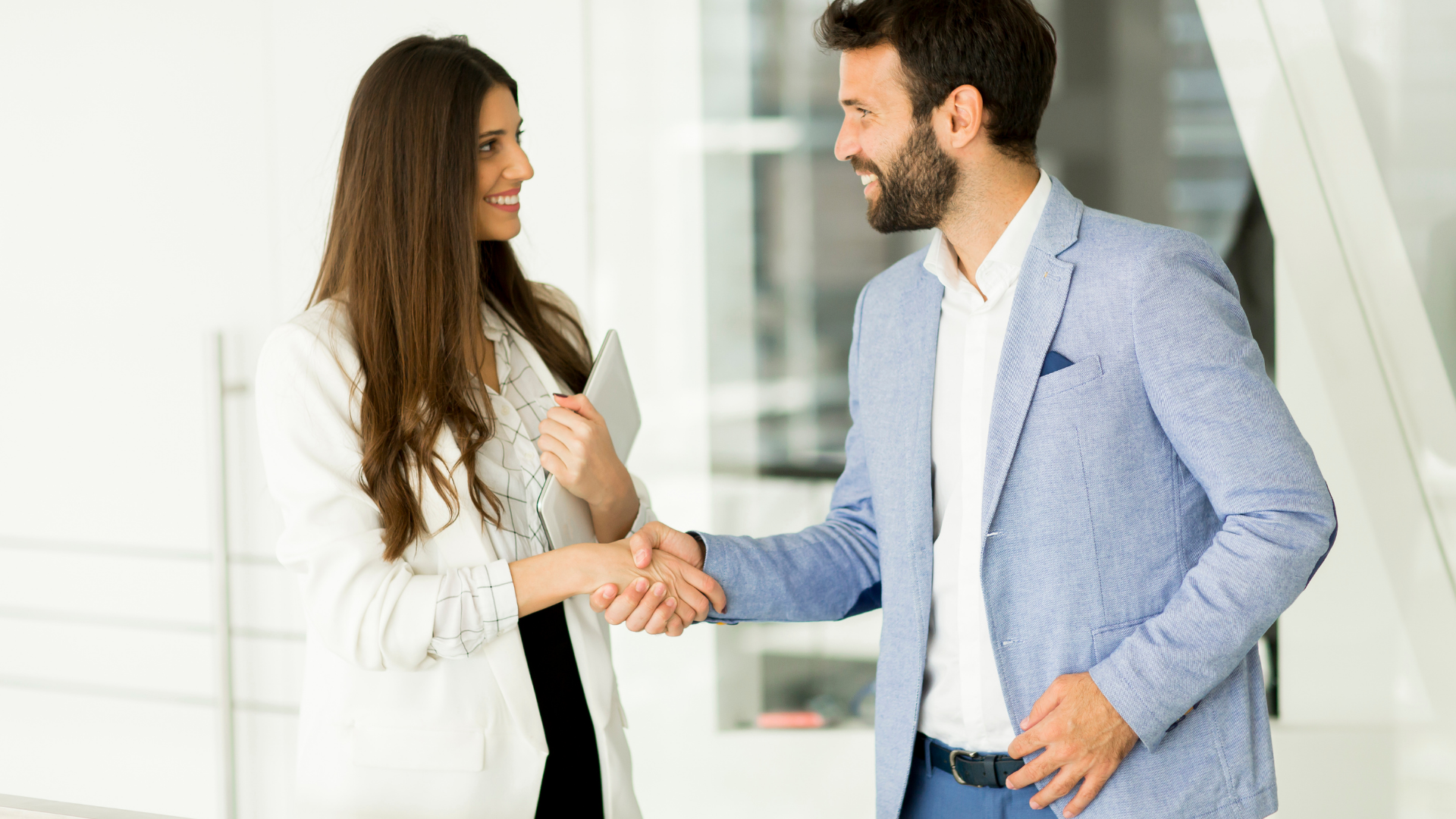 Two professionals shaking hands in a bright office, smiling and facing each other.