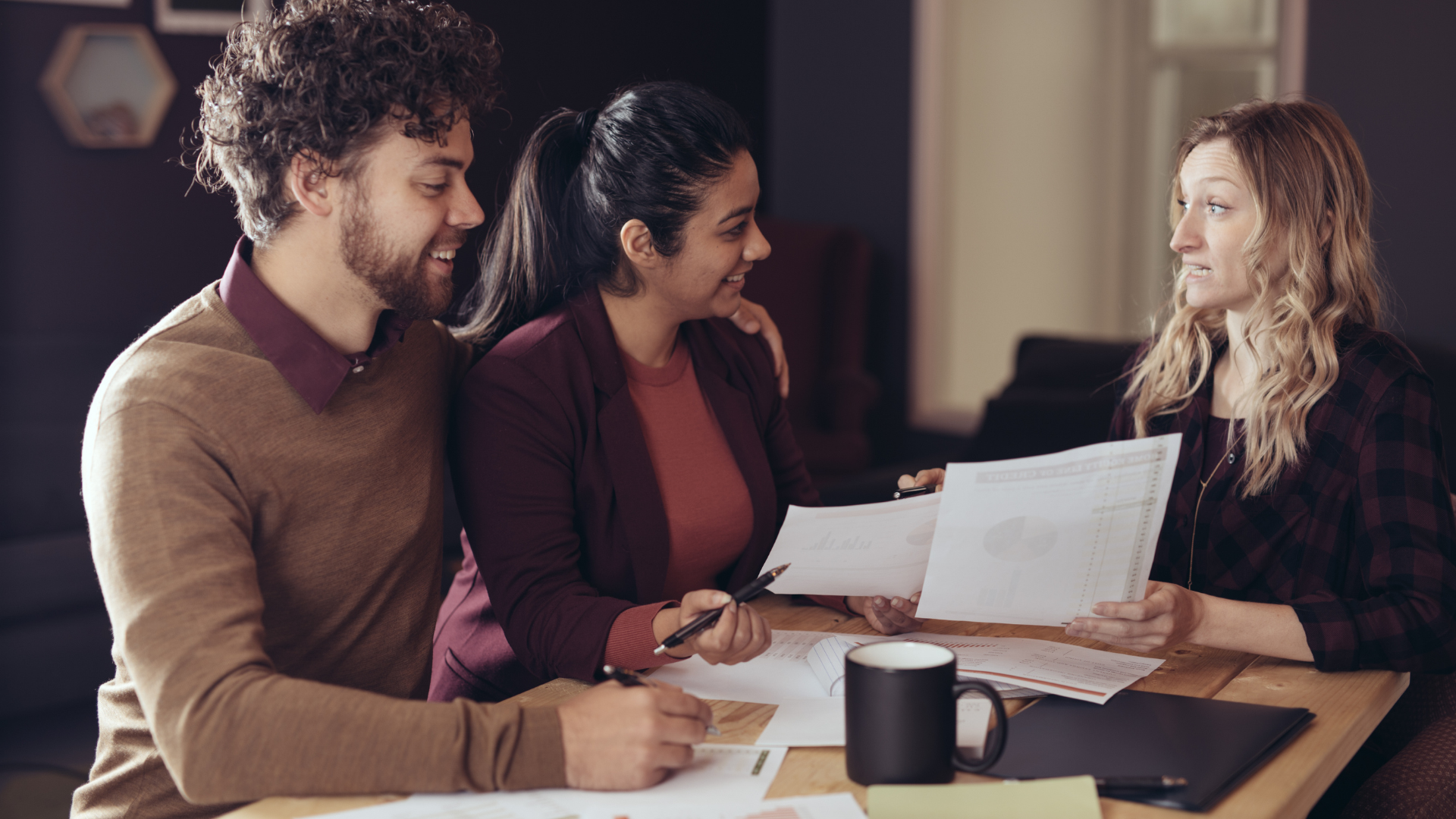 Three people review documents at a table, smiling in a warm office setting.