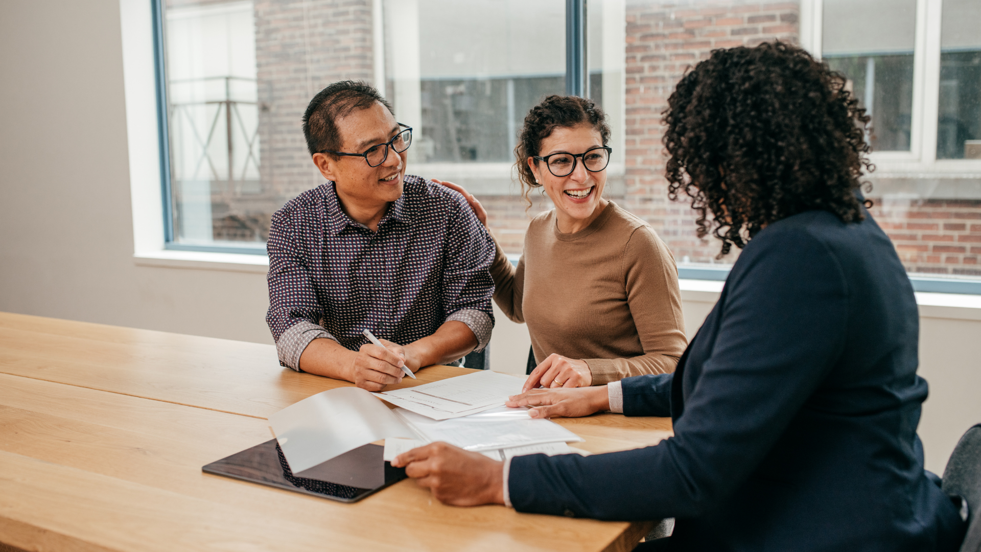 Three coworkers discuss documents at a conference table in a bright office meeting room.