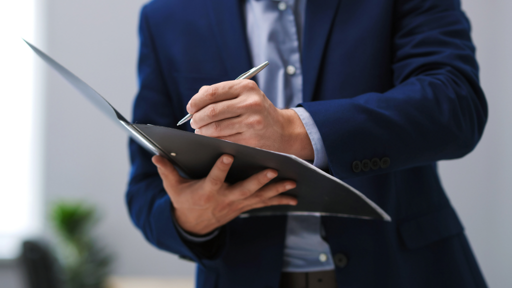 Person in a blue suit writing in a black folder, holding a pen.