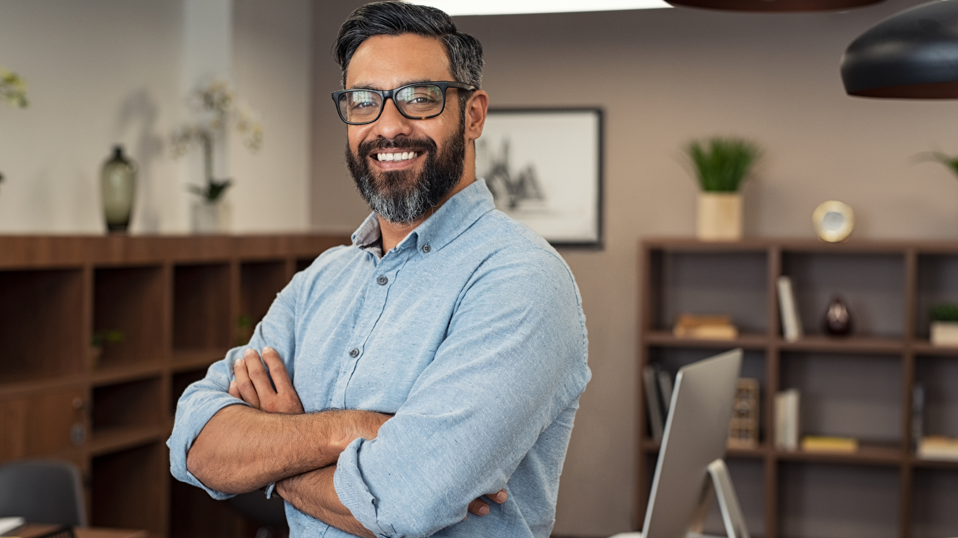 A man with glasses is standing in an office with his arms crossed and smiling.