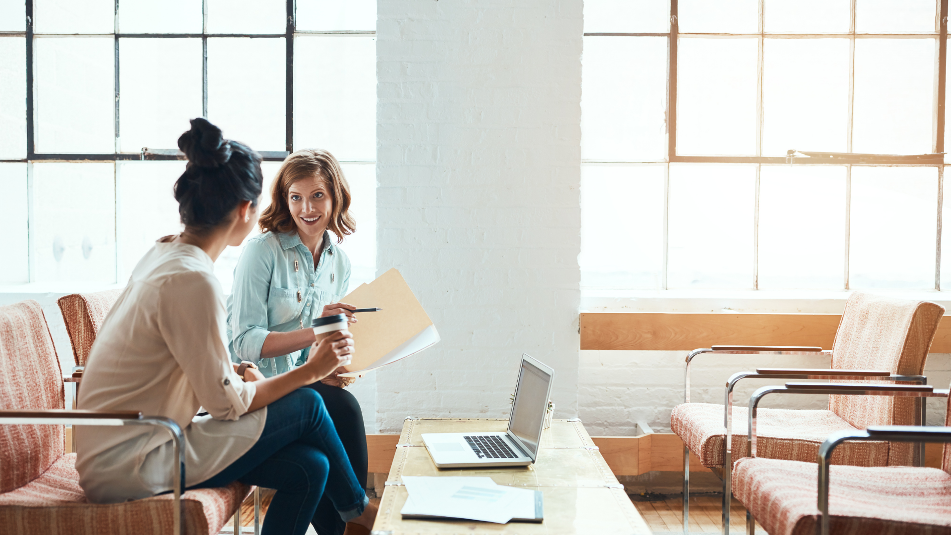 Two women in a bright office space discuss documents, one holding a coffee cup.