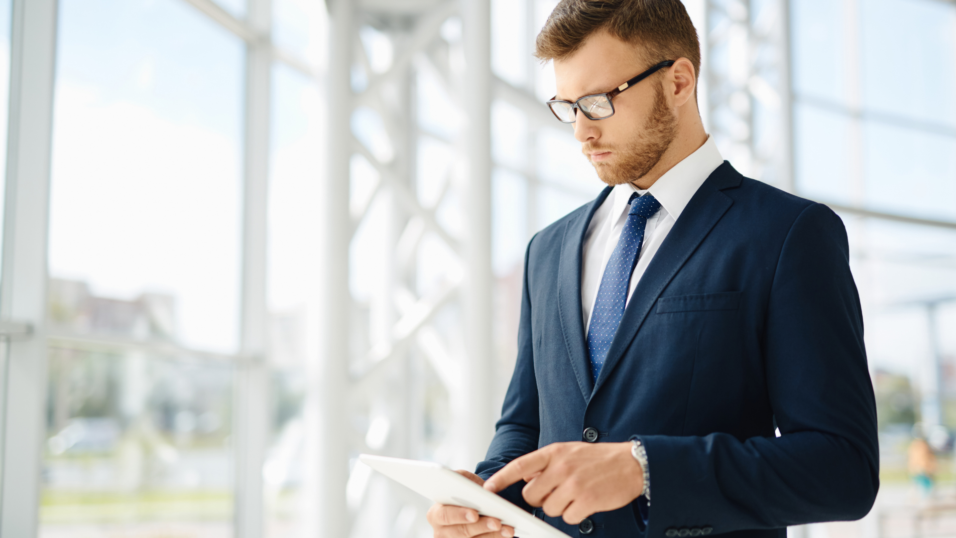 Businessman in blue suit and glasses, using a tablet near large windows.