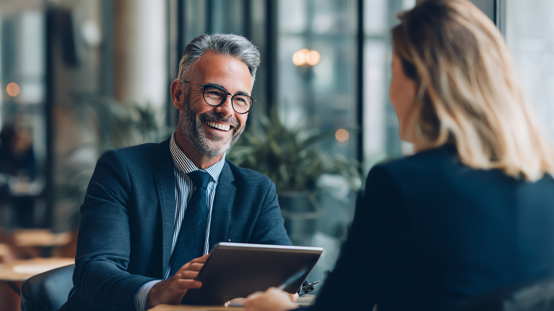 Man in suit smiles, holds tablet, speaking with another person in a modern office setting.