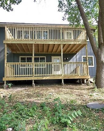 Two-story wooden deck attached to a gray house, set in a yard with grass and plants.