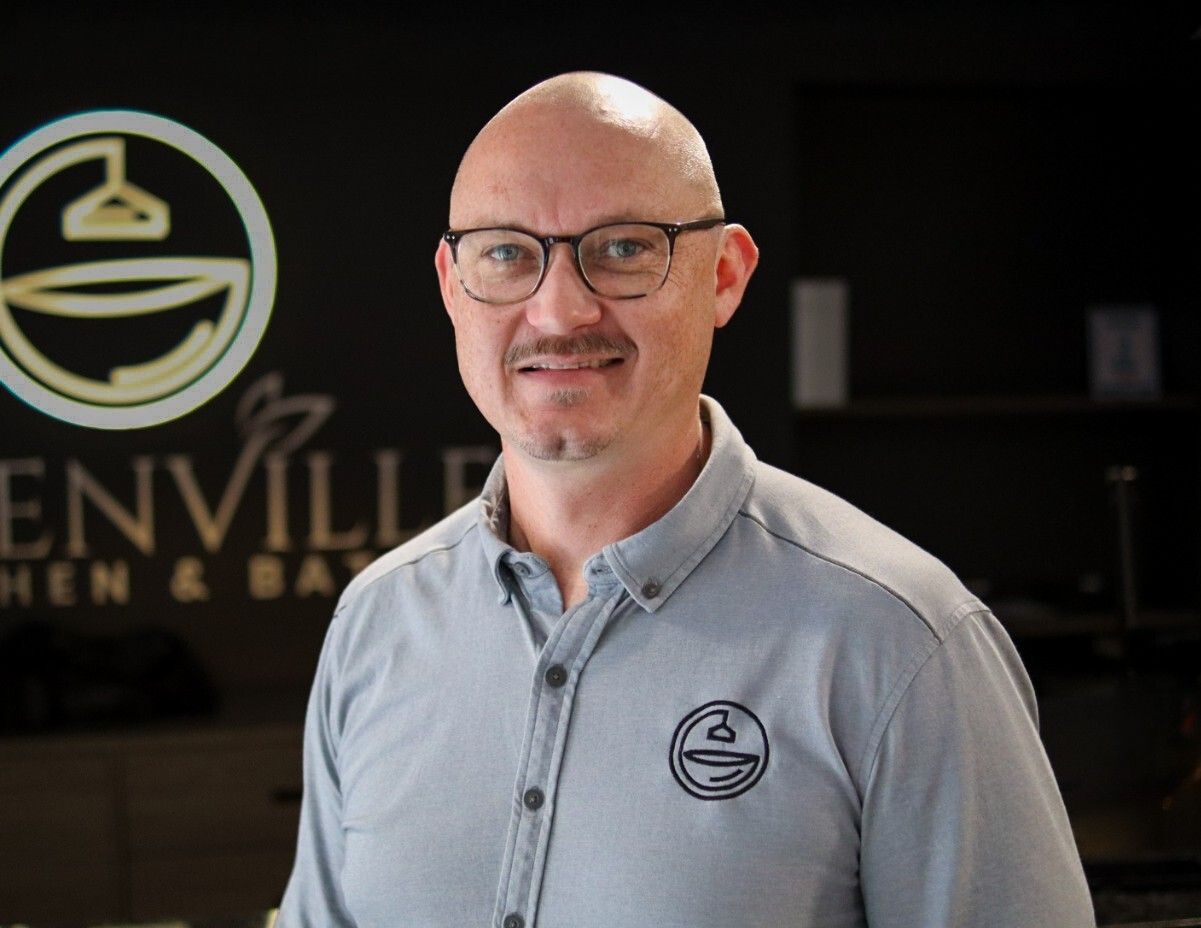 Portrait of a bald man with glasses in a gray polo, standing in front of a company logo sign.