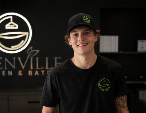 Smiling person in black shirt and cap standing in a dim café or shop with a glowing logo sign behind