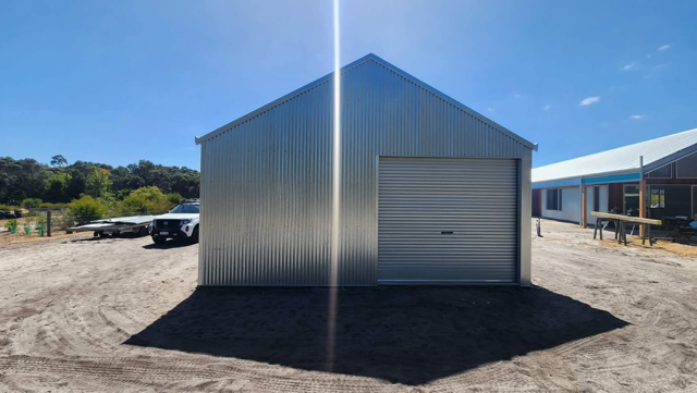 A newly constructed corrugated metal shed with a roller door stands on a gravel lot under a bright blue sky.