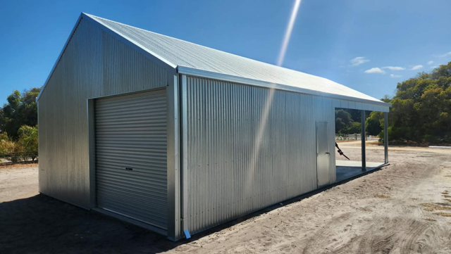 A silver, corrugated metal shed with a roller door and a covered open side, situated in a dirt yard under a sunny sky.