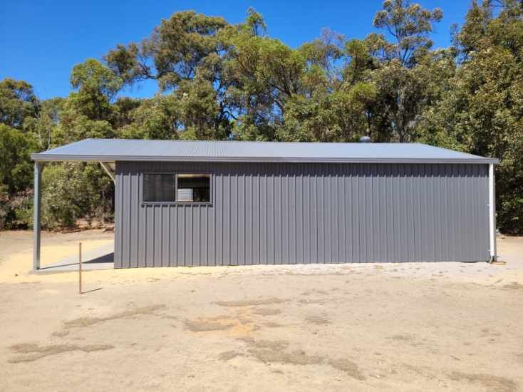 A gray metal shed with a side-roofed veranda stands in a dirt yard against a background of trees under a blue sky.