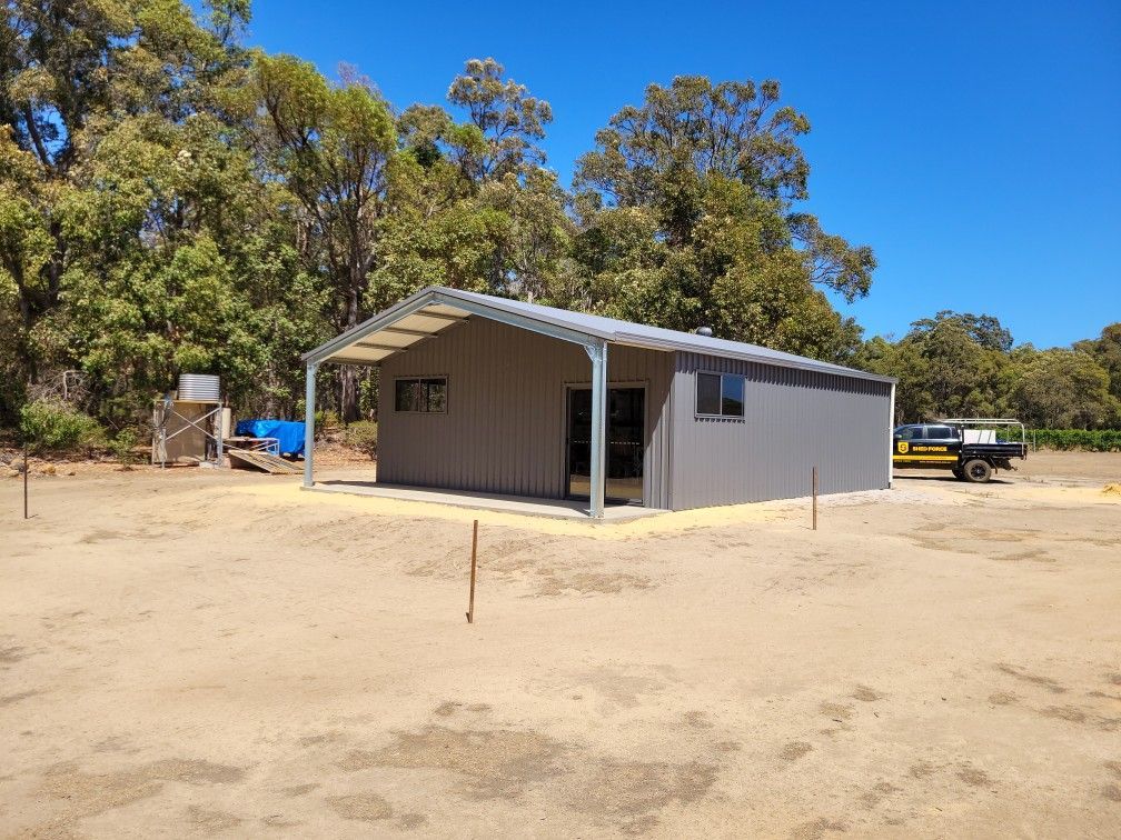 A grey metal shed with an open front and a concrete slab, set in a sunny, rural area with trees in the background.
