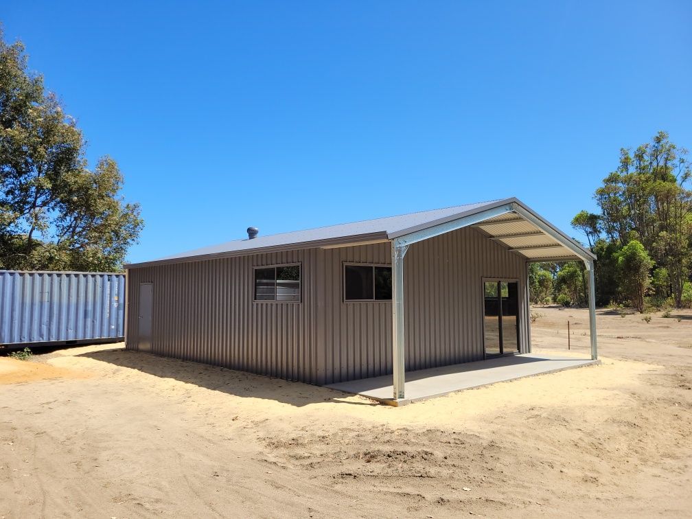 A taupe, corrugated metal shed with a side porch and windows stands on a sandy lot next to a blue shipping container.
