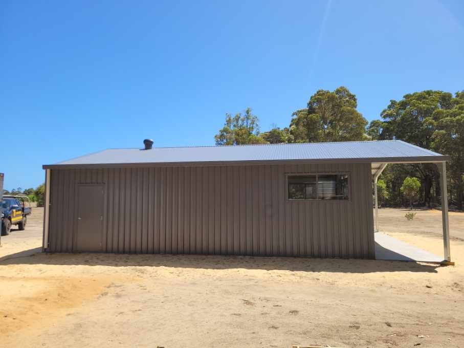 A dark gray metal shed with a matching roof, a single window, a door, and a small side porch in a sandy, outdoor setting.